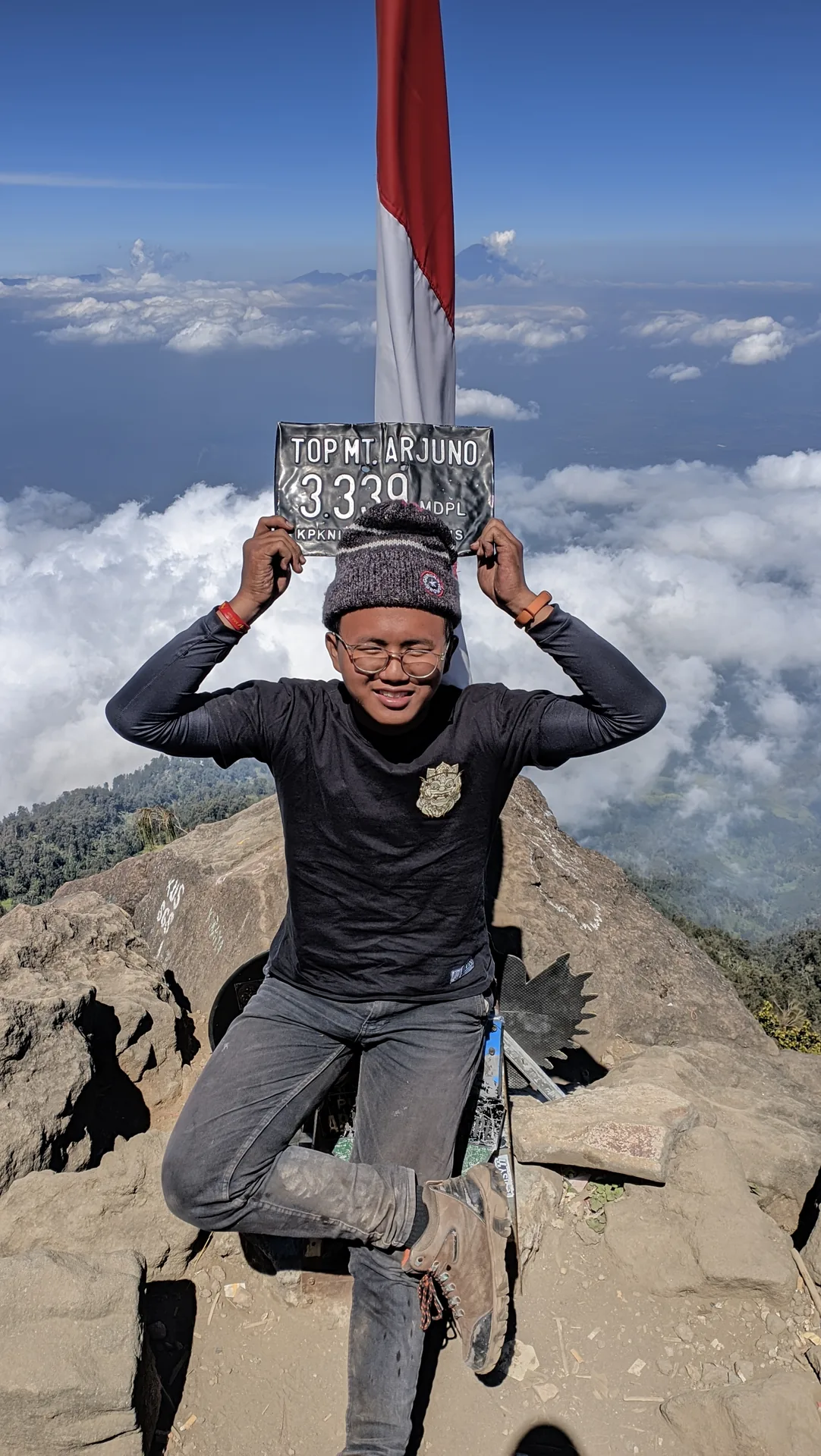 A hiker raising the Mount Arjuna summit marker above his head beneath the Indonesian flag
